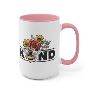 Op de afbeelding: Witte keramische mok met een roze binnenkant en handvat. De mok heeft een zwart-gele bij met bloemmotieven en het woord "KIND" in zwarte letters.