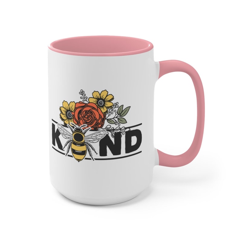 Op de afbeelding: Witte keramische mok met een roze binnenkant en handvat. De mok heeft een zwart-gele bij met bloemmotieven en het woord "KIND" in zwarte letters.