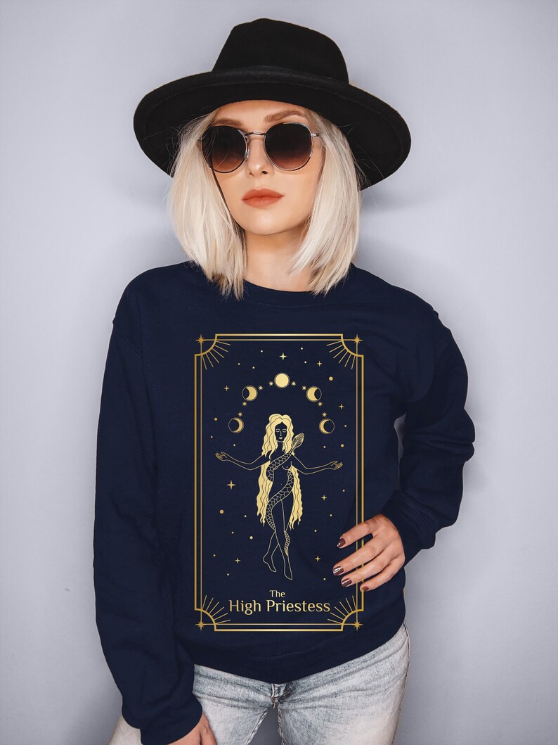Op de afbeelding: Een marineblauw sweatshirt met een gouden afbeelding van de Hogepriesteres tarotkaart. De kaart toont een vrouw met een slang om haar heen, die voor een halve maan en sterren staat.