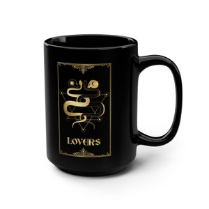 Puede incluir: Taza de caf&eacute; de cer&aacute;mica negra con un dise&ntilde;o dorado de dos serpientes entrelazadas alrededor de una calavera y un pentagrama. El texto "LOVERS" est&aacute; escrito en dorado debajo del dise&ntilde;o.