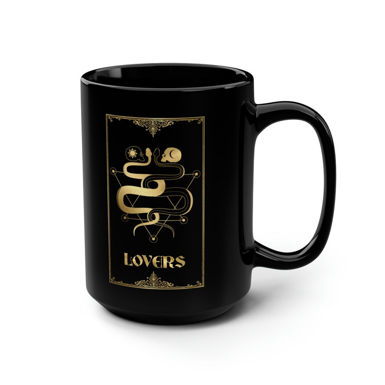 Puede incluir: Taza de caf&eacute; de cer&aacute;mica negra con un dise&ntilde;o dorado de dos serpientes entrelazadas alrededor de una calavera y un pentagrama. El texto "LOVERS" est&aacute; escrito en dorado debajo del dise&ntilde;o.