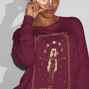 Op de afbeelding: Een bordeauxrood sweatshirt met een gouden ontwerp van de Hogepriesteres tarotkaart. De kaart toont een vrouw met een slang om haar heen, die voor een halve maan en sterren staat.