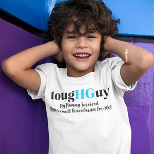 Puede incluir: Una camiseta blanca con el texto "toughGuy" en negro y azul. La camiseta también presenta la frase "My Mommy Survived Hyperemesis Gravidarum For Me!" Una persona joven lleva la camiseta y sonríe.