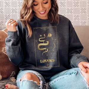 Puede incluir: Una sudadera gris oscura con un gr&aacute;fico dorado de una serpiente y la palabra "LOVERS" en la parte delantera.