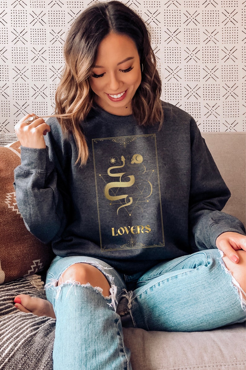Puede incluir: Una sudadera gris oscura con un gr&aacute;fico dorado de una serpiente y la palabra "LOVERS" en la parte delantera.