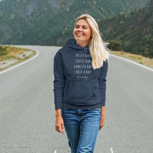 May include: A woman wearing a navy blue hoodie with the text "Messy Bun, Coffee Run, Gangsta Rap, I Need A Nap #momlife" printed on the front. She is walking on a paved road with mountains in the background.