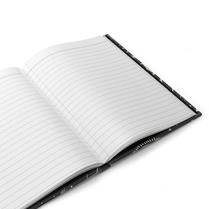 May include: An open notebook with lined pages and a black cover with a gold pattern. The notebook is lying open on a white surface.