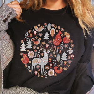 May include: Black sweatshirt with a colorful circular design featuring a deer, foxes, birds, and trees. The design is reminiscent of Scandinavian folk art.