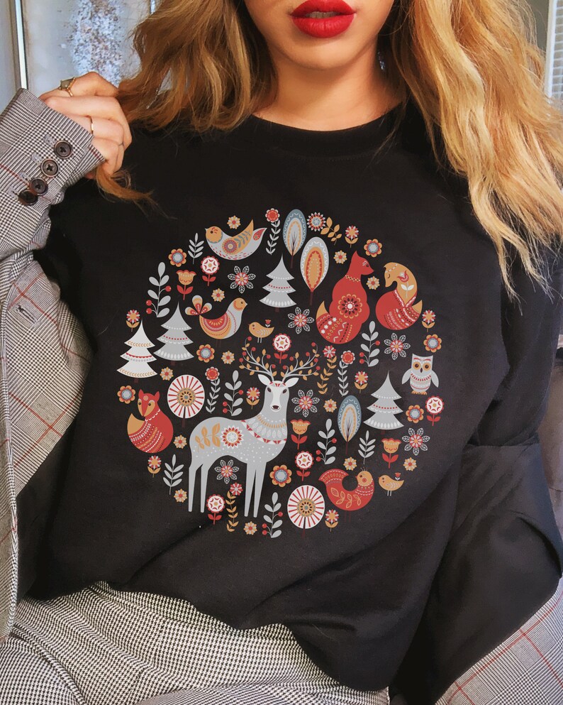 May include: Black sweatshirt with a colorful circular design featuring a deer, foxes, birds, and trees. The design is reminiscent of Scandinavian folk art.