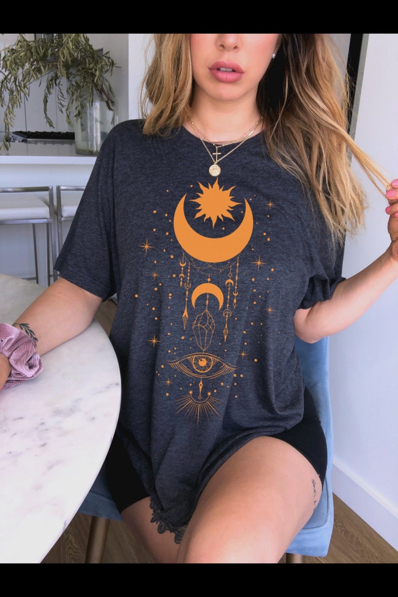 May include: A dark gray t-shirt with an orange celestial design featuring a crescent moon, sun, stars, and an eye with a crystal.