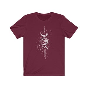 May include: A maroon t-shirt with a white graphic of three moons and stars. The moons are in different phases and are connected by a line of stars.