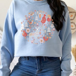 May include: A light blue sweatshirt with a circular design featuring a deer, fox, birds, and trees in a Scandinavian folk art style.
