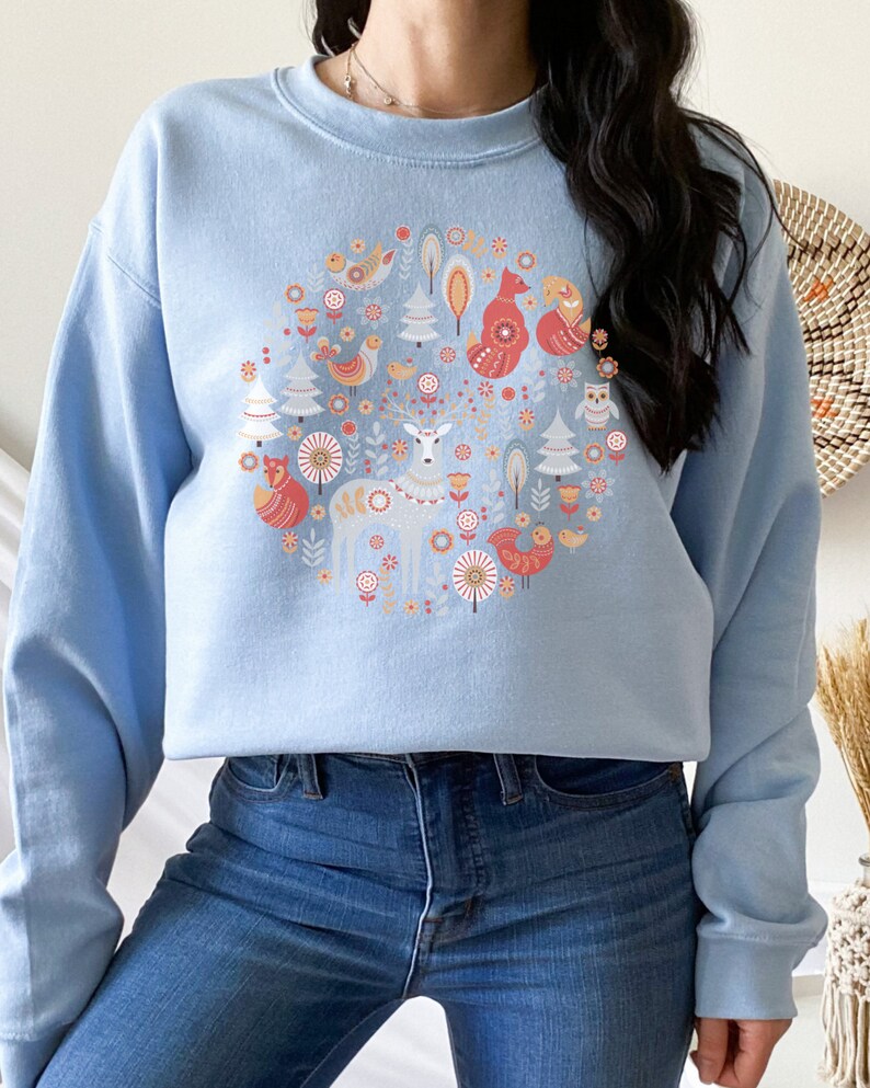 May include: A light blue sweatshirt with a circular design featuring a deer, fox, birds, and trees in a Scandinavian folk art style.