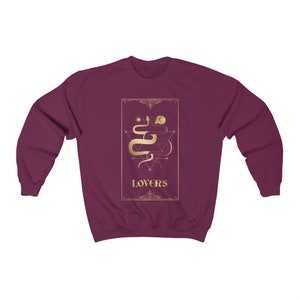 Puede incluir: Sudadera color burdeos con un gr&aacute;fico dorado de una serpiente y formas geom&eacute;tricas. El gr&aacute;fico est&aacute; rodeado por un marco dorado y la palabra "LOVERS" est&aacute; escrita debajo del gr&aacute;fico.