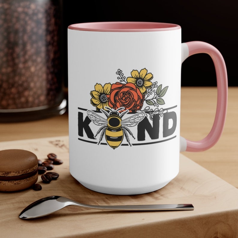 Op de afbeelding: Een witte keramische mok met een roze rand en handvat. De mok heeft een bij met gele en zwarte strepen, omringd door bloemen in tinten geel, oranje en rood. Het woord "KIND" is in zwarte letters onder de bij en de bloemen gedrukt.