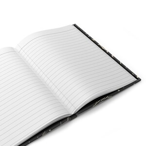 May include: An open notebook with lined pages and a black cover with a gold pattern. The notebook is lying open on a white surface.