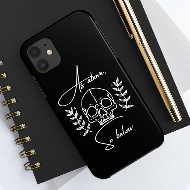 Op de afbeelding: Zwarte telefoonhoes met een wit schedel- en bladerenontwerp. De tekst "As above, so below" is geschreven in een cursief schrift.