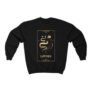 Puede incluir: Sudadera negra con un dise&ntilde;o dorado de una serpiente con un sol y una luna encima. El dise&ntilde;o est&aacute; en un marco con la palabra "LOVERS" debajo.