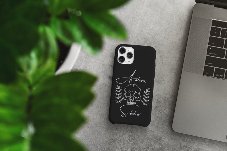 Op de afbeelding: Zwarte telefoonhoes met een witte schedel en de tekst "As above, so below".