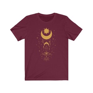 May include: A maroon t-shirt with a gold design of a crescent moon, sun, eye, and other celestial symbols.