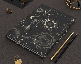 May include: A black notebook with a celestial design featuring moons, stars, and sun symbols in a cream color. The notebook is lying on a dark gray textured surface with gold paper clips and pencils.