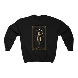 Op de afbeelding: Zwart sweatshirt met een gouden ontwerp van de Hogepriesteres tarotkaart. De kaart toont een vrouw die met haar armen gespreid staat, omringd door manen en sterren.