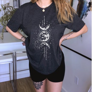 May include: A dark gray t-shirt with a white moon and stars graphic. The shirt has a relaxed fit and a round neckline.