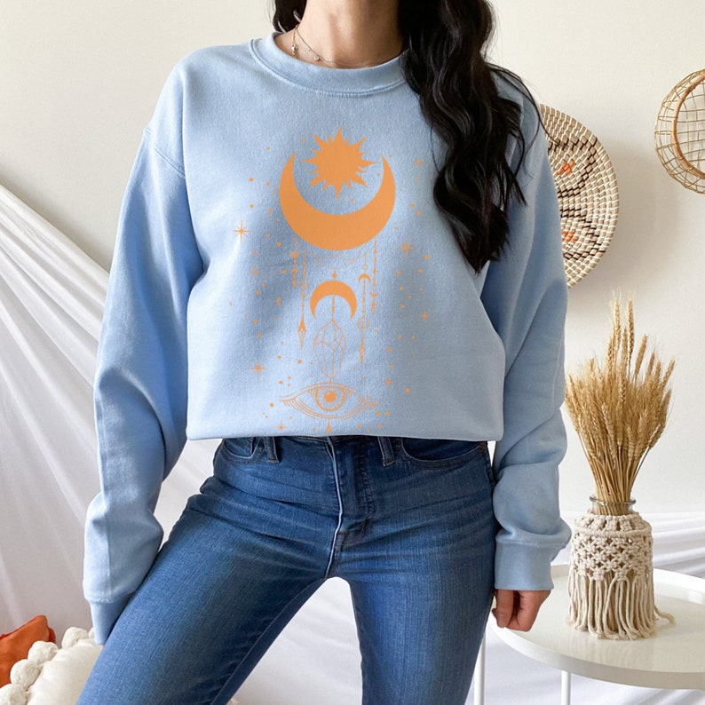 May include: A light blue sweatshirt with a graphic design of a crescent moon, a star, and an eye. The design is in a peach color.