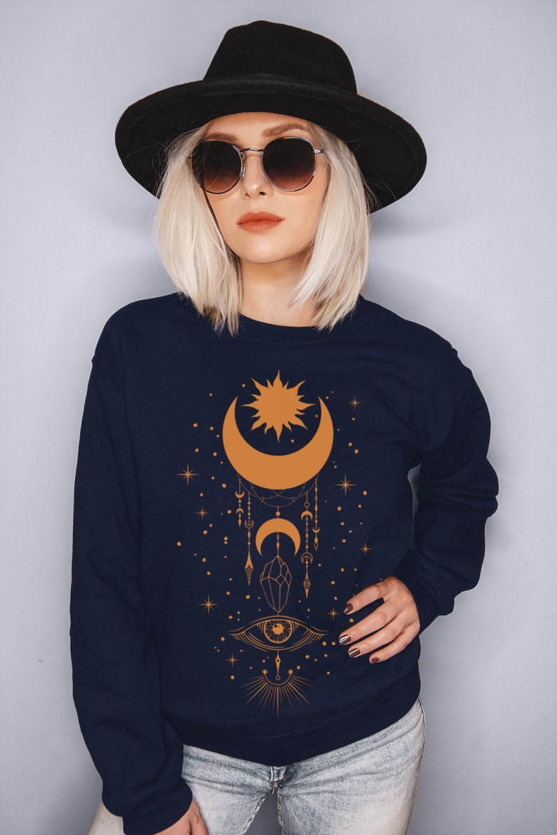May include: A navy blue sweatshirt with a gold sun and moon design. The design features a crescent moon with a sun inside, a crystal, and an eye with rays of light. The sweatshirt has a relaxed fit and is perfect for casual wear.