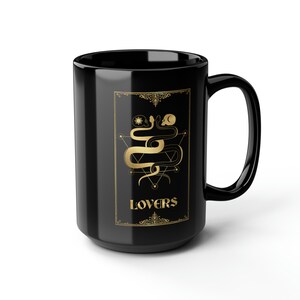 Puede incluir: Taza de caf&eacute; de cer&aacute;mica negra con un dise&ntilde;o dorado de dos serpientes entrelazadas alrededor de un s&iacute;mbolo geom&eacute;trico. El texto "LOVERS" est&aacute; escrito en dorado debajo de las serpientes.