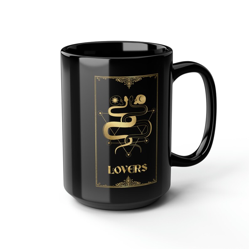 Puede incluir: Taza de caf&eacute; de cer&aacute;mica negra con un dise&ntilde;o dorado de dos serpientes entrelazadas alrededor de un s&iacute;mbolo geom&eacute;trico. El texto "LOVERS" est&aacute; escrito en dorado debajo de las serpientes.