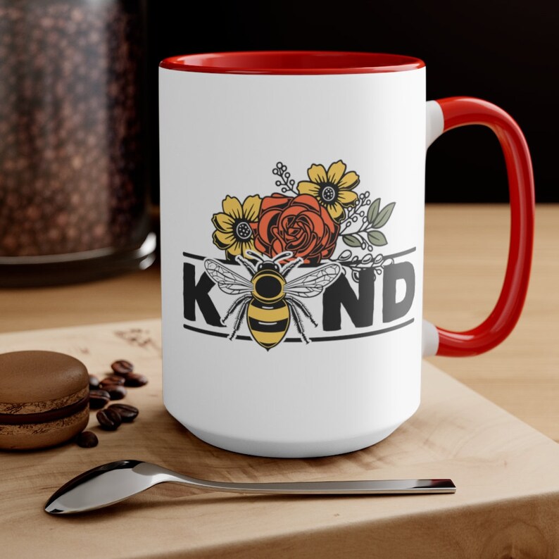 Op de afbeelding: Witte keramische mok met een rode rand en handvat. De mok heeft een afbeelding van een bij met bloemen en het woord "KIND" in zwarte letters.
