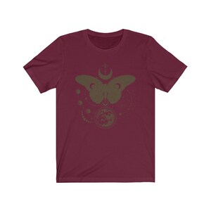 May include: A maroon short-sleeved t-shirt with a graphic print of a butterfly with a crescent moon and stars. The butterfly is in a dark green color.