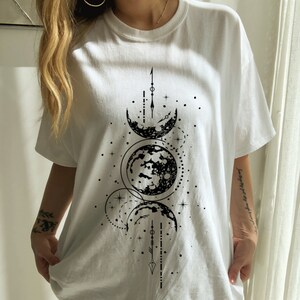 May include: White t-shirt featuring a black graphic of the moon phases. The design includes crescent moons, a full moon, stars, and geometric shapes. The shirt is a casual, oversized fit.