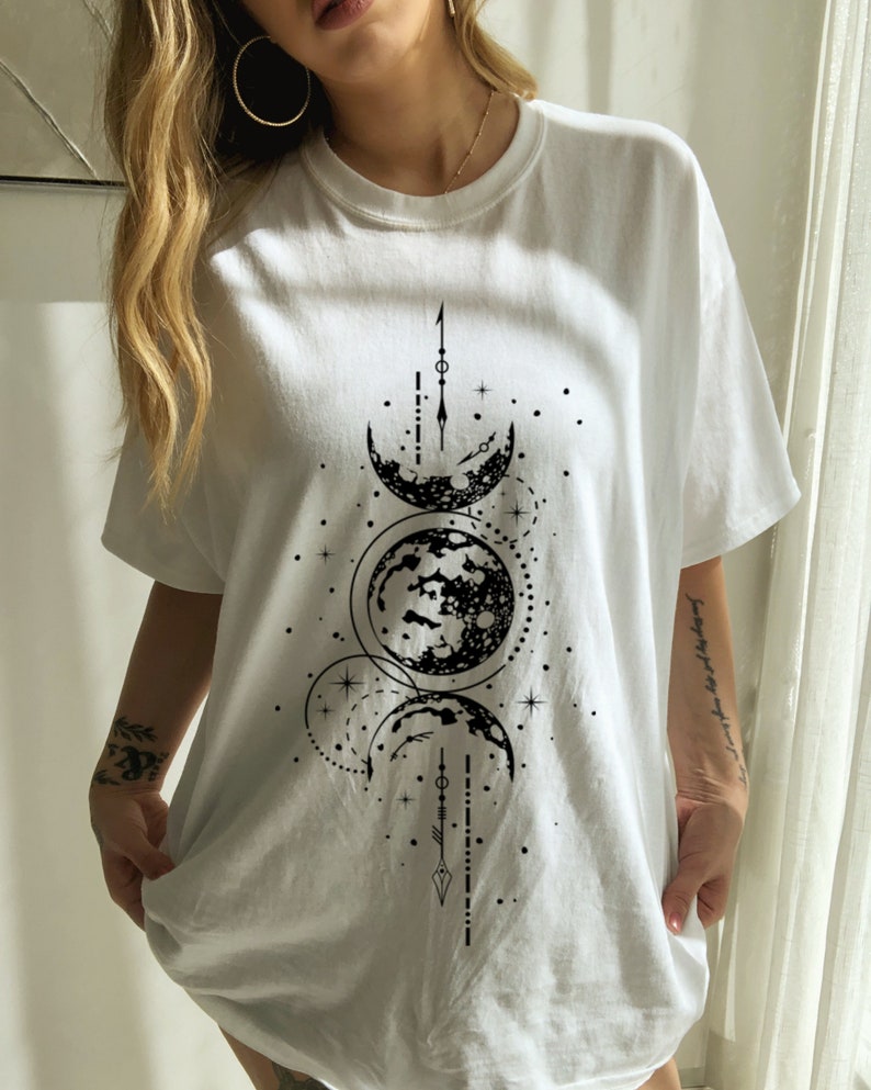 May include: White t-shirt featuring a black graphic of the moon phases. The design includes crescent moons, a full moon, stars, and geometric shapes. The shirt is a casual, oversized fit.