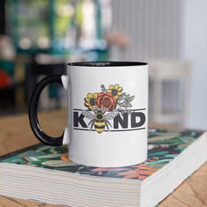 Op de afbeelding: Witte keramische mok met een zwarte handgreep. De mok heeft een zwarte rand en heeft een bij met bloemen en het woord "KIND" in zwarte letters.