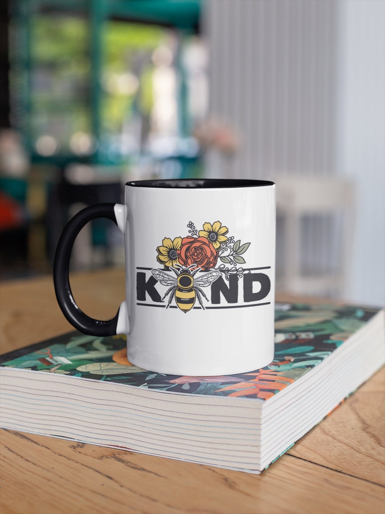 Op de afbeelding: Witte keramische mok met een zwarte handgreep. De mok heeft een zwarte rand en heeft een bij met bloemen en het woord "KIND" in zwarte letters.