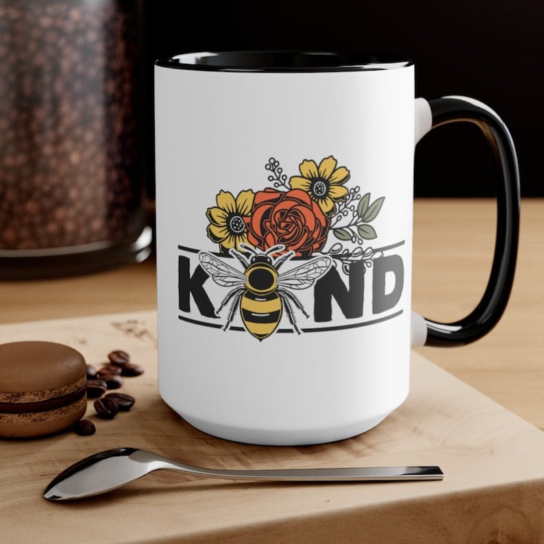 Op de afbeelding: Een witte keramische mok met een zwarte rand en handvat. De mok heeft een bij met gele en zwarte strepen, omringd door bloemen in tinten rood, geel en groen. Het woord "KIND" is in zwarte letters boven de bij gedrukt.