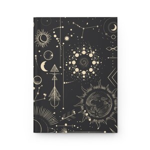 May include: A black notebook cover with a white celestial pattern. The pattern includes stars, moons, suns, and geometric shapes.