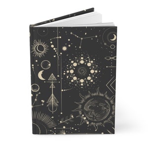 May include: A black journal with a gold celestial pattern. The pattern includes moons, stars, suns, and geometric shapes.