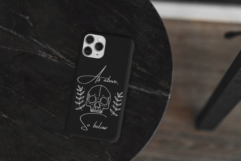 Op de afbeelding: Zwarte telefoonhoes met een witte schedel en een bloemenontwerp. De tekst "As above, so below" is op de hoes gedrukt.