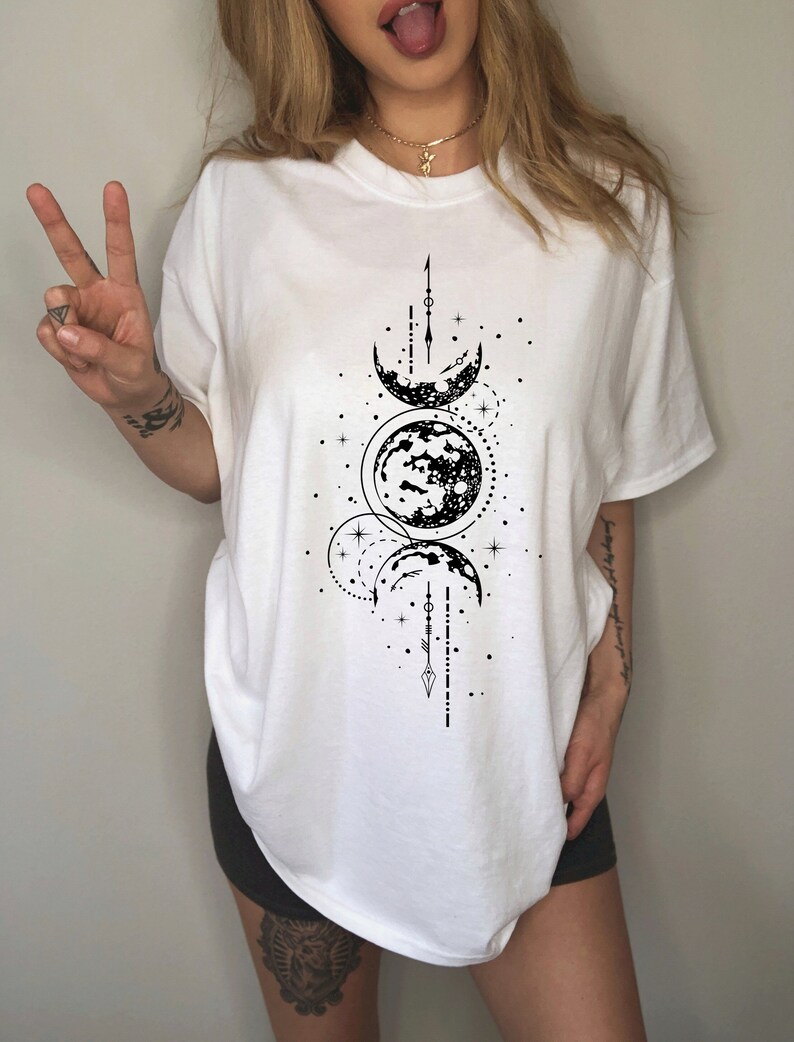 May include: White t-shirt featuring a black graphic of moon phases and celestial elements. The design includes crescent moons, full moons, stars, and linear details. The shirt has a crew neck and short sleeves.