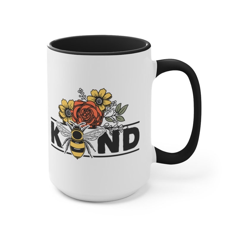 Op de afbeelding: Een witte keramische mok met een zwarte rand en handvat. De mok heeft een bij met gele en zwarte strepen, omringd door bloemen in tinten rood, geel en oranje. Het woord "KIND" is in zwarte letters onder de bij en bloemen gedrukt.