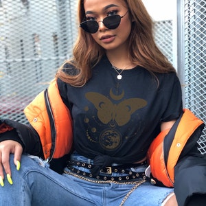 May include: A black t-shirt with a gold butterfly design and celestial elements. The butterfly has a crescent moon on its back and is surrounded by stars and planets. The shirt is worn with a black belt and a gold chain.