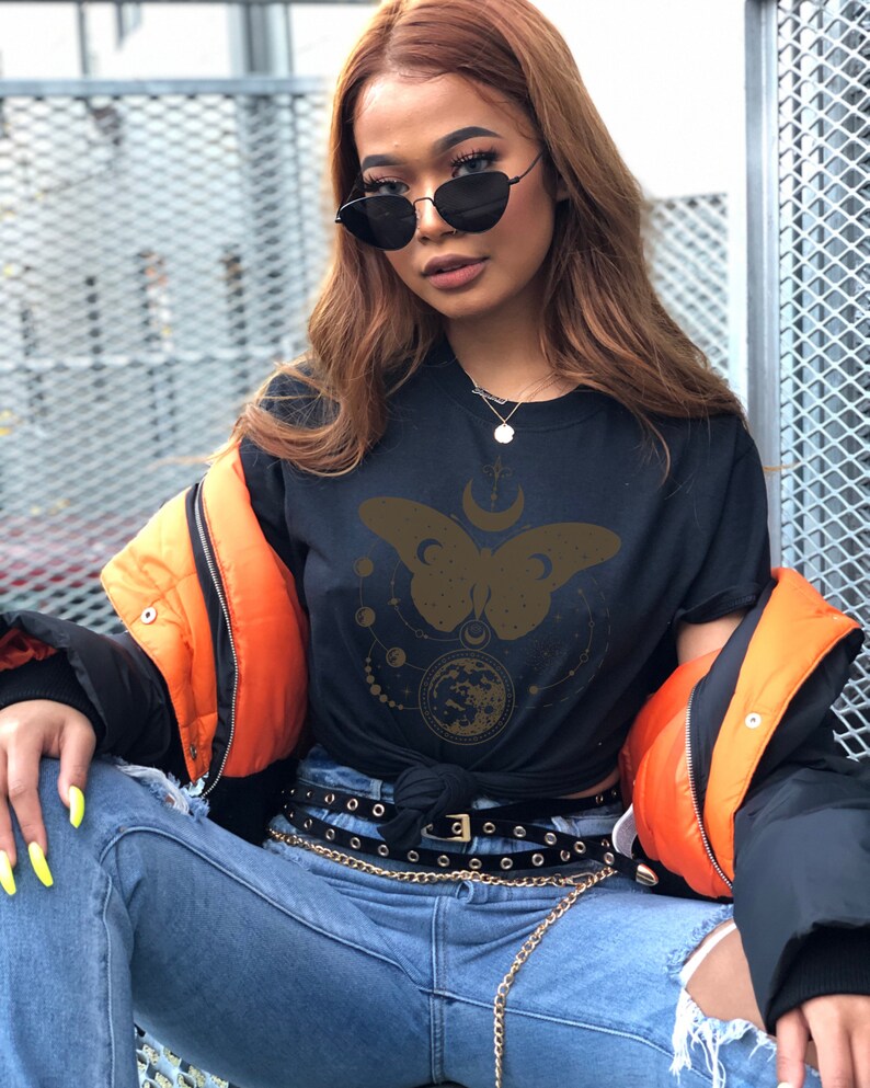 May include: A black t-shirt with a gold butterfly design and celestial elements. The butterfly has a crescent moon on its back and is surrounded by stars and planets. The shirt is worn with a black belt and a gold chain.