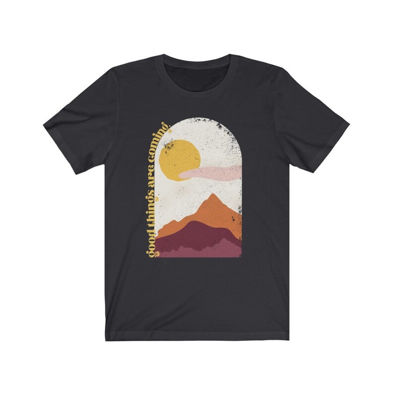 Puede incluir: Camiseta negra con un gr&aacute;fico de un sol, monta&ntilde;as y una mano que se estira hacia arriba. El texto "good things are coming" est&aacute; escrito verticalmente en el lado izquierdo del gr&aacute;fico.