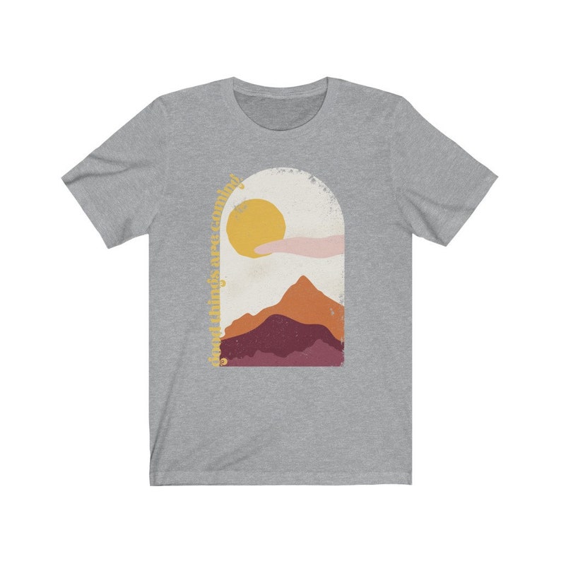 Puede incluir: Camiseta gris con un gr&aacute;fico de una cordillera, un sol y una nube. El texto "Good things are coming" est&aacute; escrito en una fuente de letra cursiva alrededor del gr&aacute;fico.