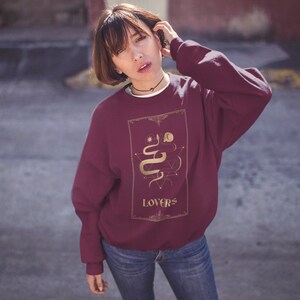 Puede incluir: Una sudadera color burdeos con un gr&aacute;fico dorado de una serpiente y la palabra "LOVERS" en la parte delantera.