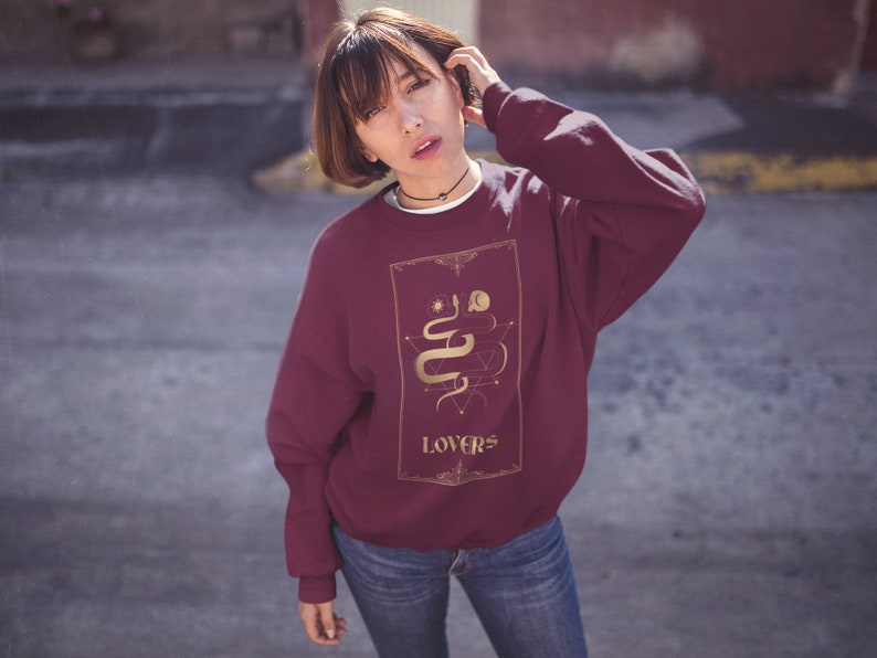 Puede incluir: Una sudadera color burdeos con un gr&aacute;fico dorado de una serpiente y la palabra "LOVERS" en la parte delantera.