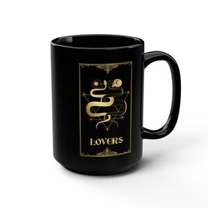 Puede incluir: Taza de caf&eacute; de cer&aacute;mica negra con un dise&ntilde;o dorado de dos serpientes entrelazadas alrededor de un pentagrama con la palabra "LOVERS" en letras doradas.
